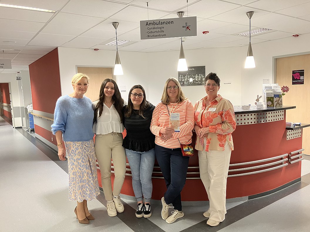 Fünf Frauen stehen nebeneinander in einem Krankenhausflur vor einem Empfangstresen mit Schild 'Ambulanzen'