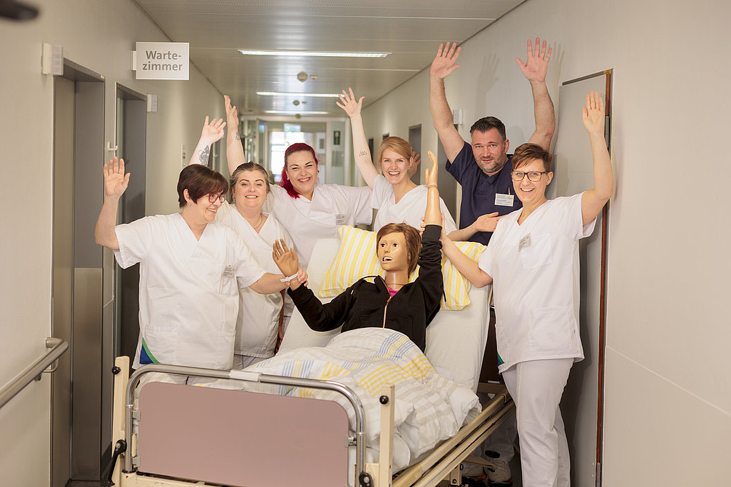 Krankenhauspersonal in weißen Kitteln hebt im Flur die Hände, Patient im Bett mit erhobenen Armen